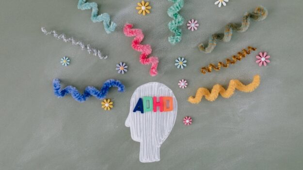 adhd Illustratie van een hoofd met de letters "ADHD" erop, omringd door kleurrijke kronkelige pijpenragers en bloemen, die de chaos en prikkels in het brein symboliseren.
