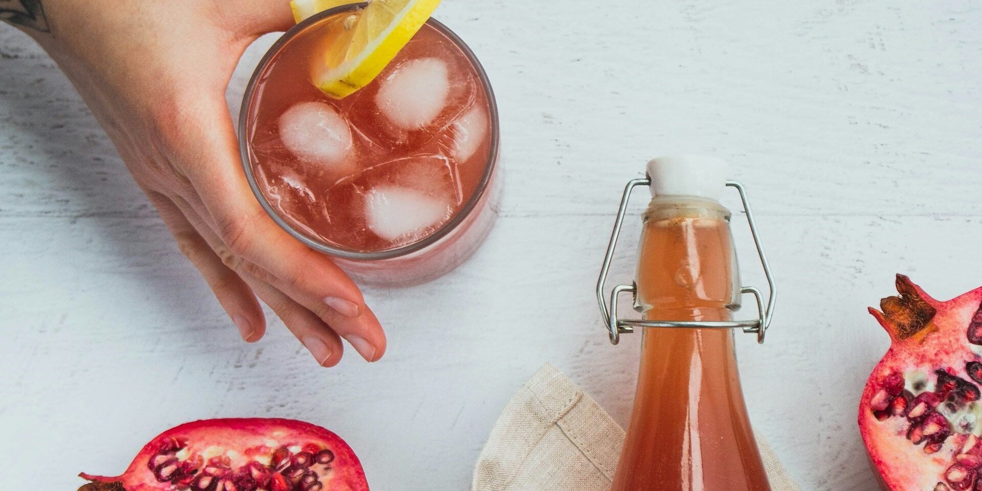 Verfrissende kombucha met granaatappel en citroen Glas kombucha met ijs en citroen, naast een fles en halve granaatappels op een lichte ondergrond.