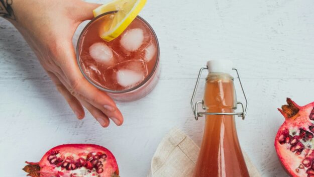 Verfrissende kombucha met granaatappel en citroen Glas kombucha met ijs en citroen, naast een fles en halve granaatappels op een lichte ondergrond.