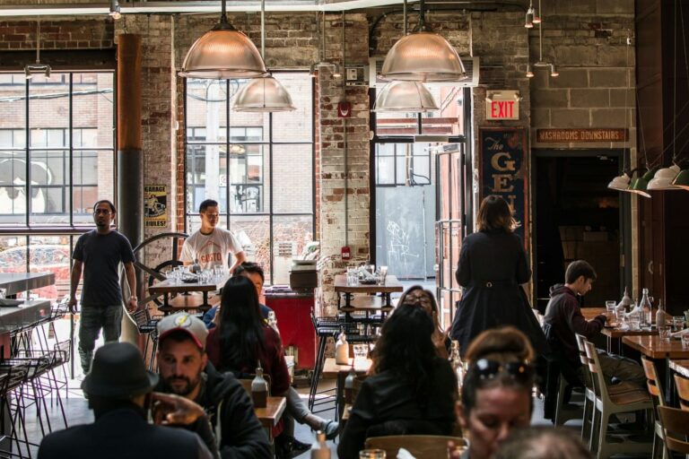 Binnenkant van een restaurant met een industriële uitstraling, featuring grote ramen, zichtbare bakstenen muren, en mensen die zowel als klanten als personeel bewegen. Het restaurant heeft een moderne en open sfeer.